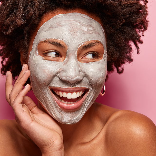 Photo of glad black woman has clay mask, enjoys facial treatments, keeps gaze aside, touches face, has spa therapy, broad smile with white teeth, isolated over pink background. Perfect skin concept